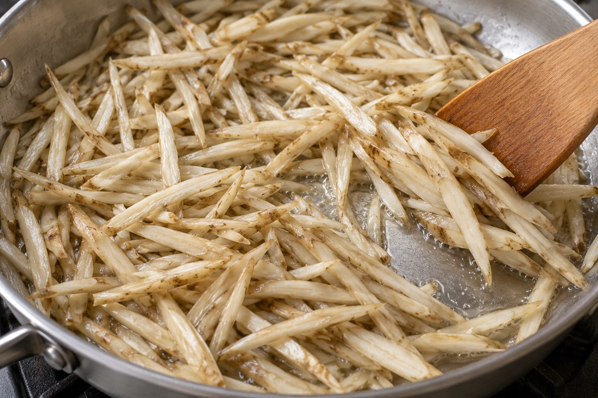 Pale burdock strips and shallow liquid in a pan with a wooden spatula