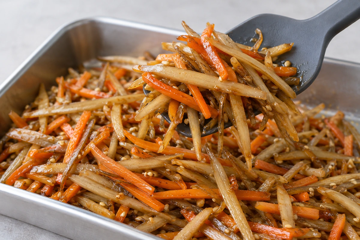 Wide spatula lifting cooked burdock and carrot strips from a stainless tray Wide spatula lifting cooked burdock and carrot strips from a stainless tray